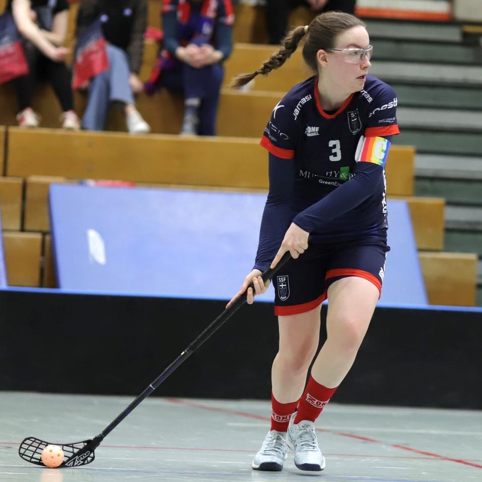 Sportlerin in blauem Trikot mit Nummer 3 dribbelt einen Floorball auf dem Spielfeld in einer Sporthalle.