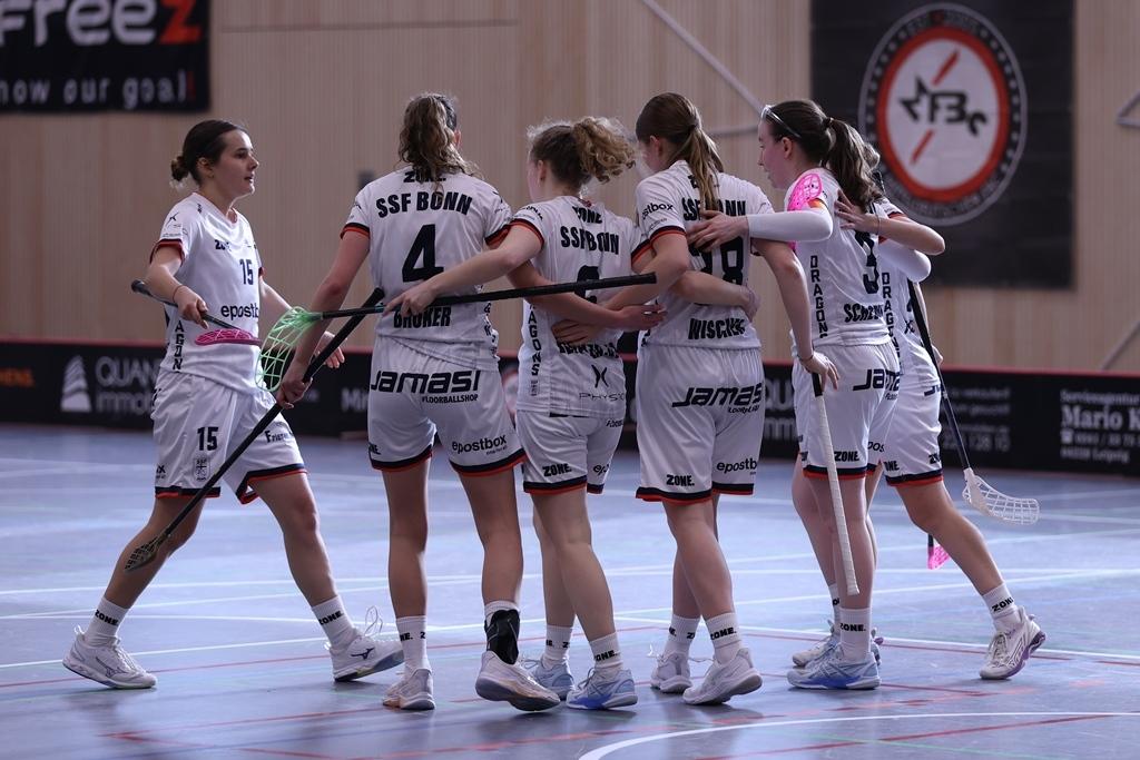 Sechs Frauen in Sportkleidung umarmen sich glücklich auf einem Hallenboden nach einem Spiel.