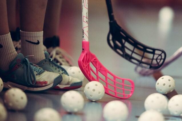 Nahaufnahme von Füßen in Sportschuhen und zwei Floorball-Schlägern, umgeben von weißen Ballen auf dem Boden.
