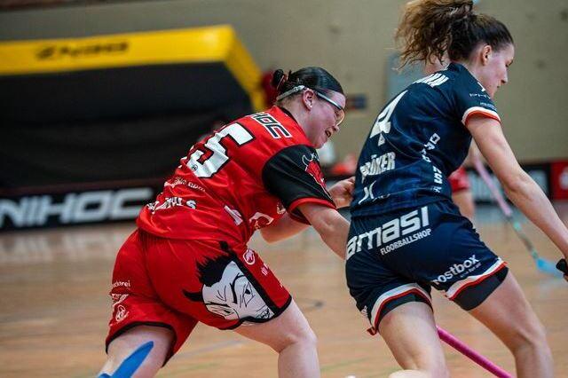 Zwei Frauen in Sportkleidung laufen in einem Hallenboden mit Schlägern in den Händen aufeinander zu.
