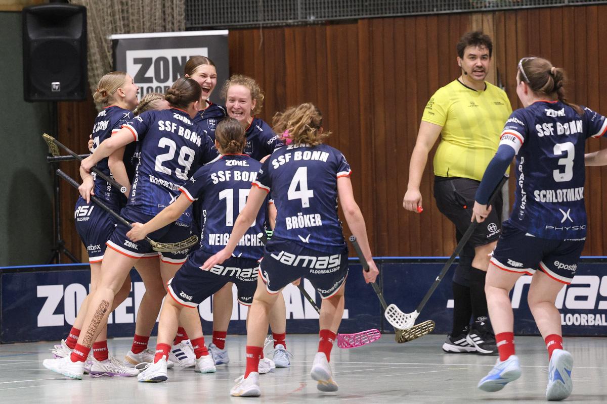 Acht Frauenfeiern im Teamtrikot von SSF Bonn, während ein Schiedsrichter neben ihnen steht.