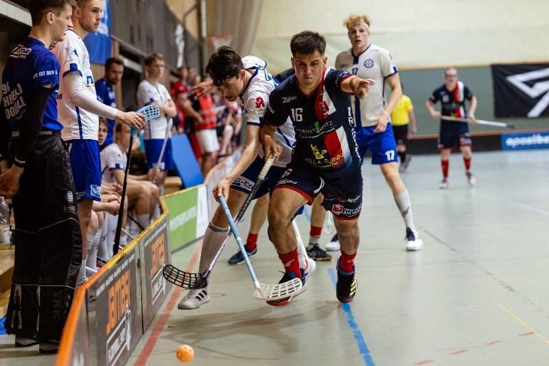Zwei Spieler kämpfen um den Ball in einem Hallenboden-Hockeyspiel, während Zuschauer auf der Bank zuschauen.
