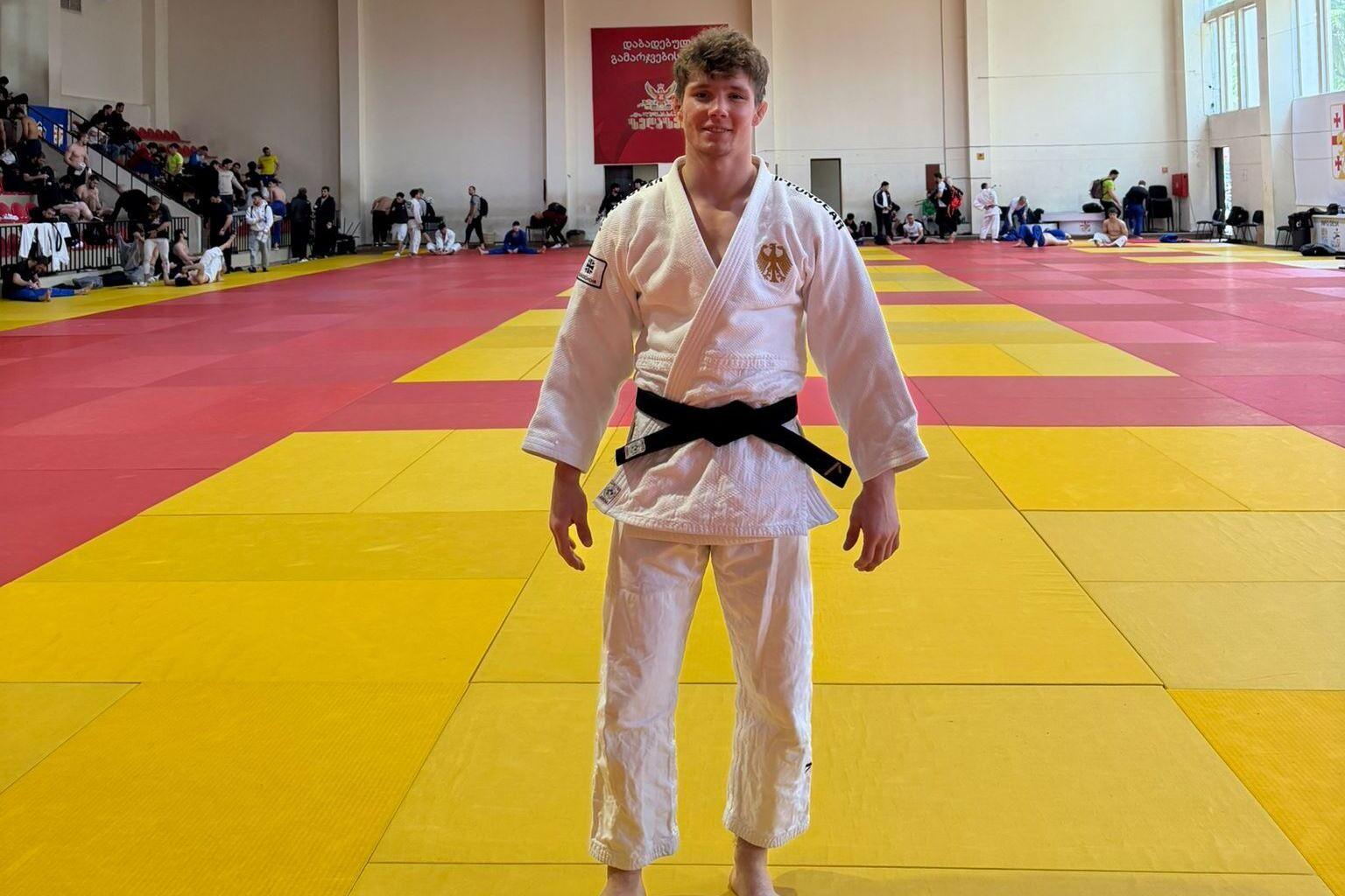 Junger Judoka in weißem Gi mit schwarzem Gürtel auf einer bunten Judomatte, im Hintergrund sind andere Kämpfer und Zuschauer.