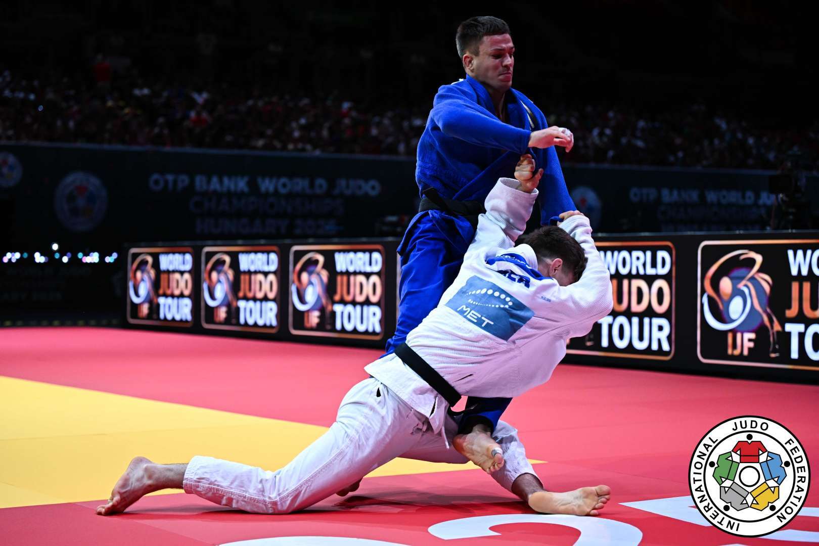 Zwei Judokämpfer in Aktion auf einer rot-gelben Matte während eines Wettkampfs. Der Kämpfer in Blau greift den Gegner in Weiß an.