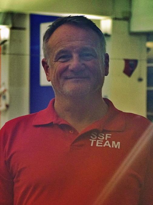 Mann mit grau melierten Haaren und freundlichem Lächeln in rotem Shirt mit „SSF TEAM“ vor einem Schwimmbecken.