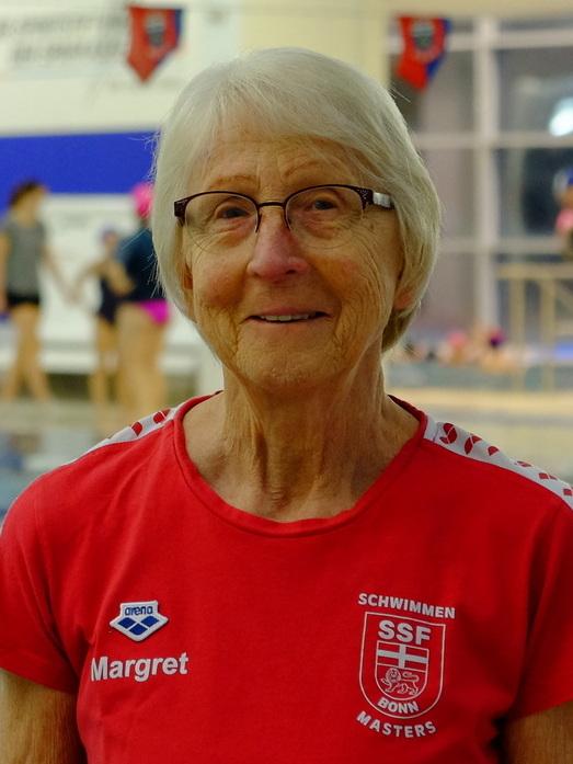 Ältere Schwimmerin mit Brille in rotem Shirt der SSF Bonn, Hintergrund mit Schwimmbecken und Übenden.