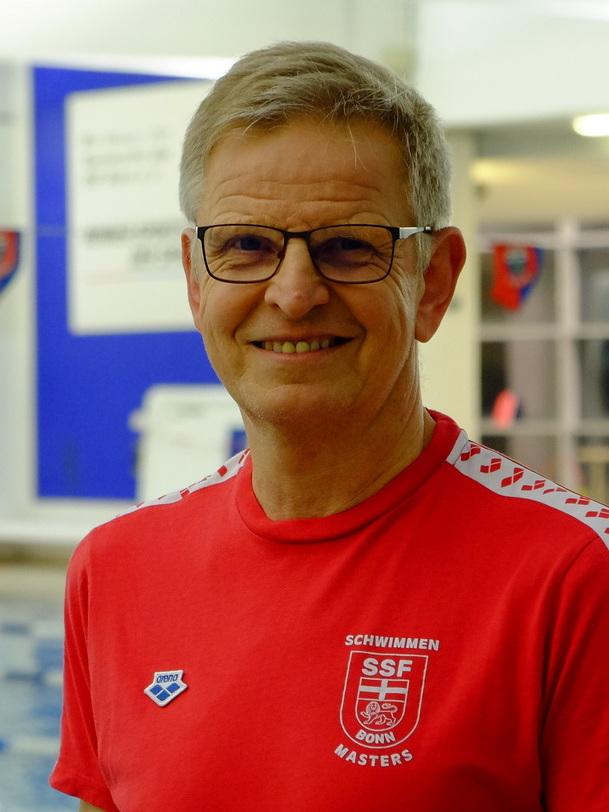 Lächelnder Mann in rotem T-Shirt mit Schwimmen-Logo steht am Beckenrand eines Schwimmbads.