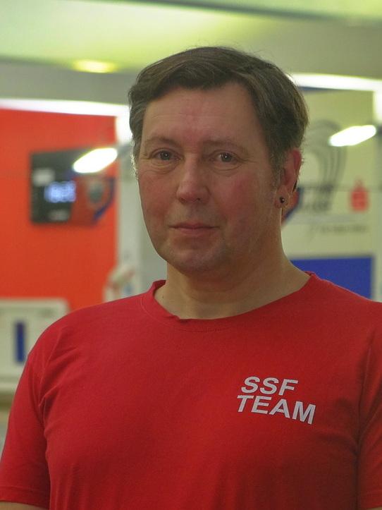 Lächelnder Mann in rotem T-Shirt mit „SSF TEAM“-Aufdruck, stehend in einer Schwimmhalle im Hintergrund.