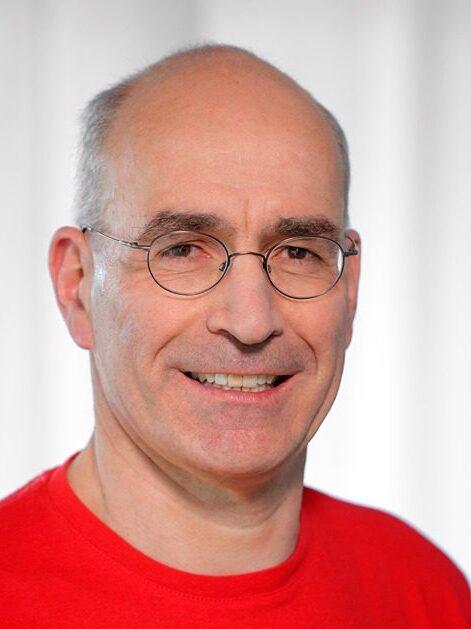 Porträt eines Mannes mit Brille, der in einem roten Shirt lächelt, vor hellem, verschwommenem Hintergrund.
