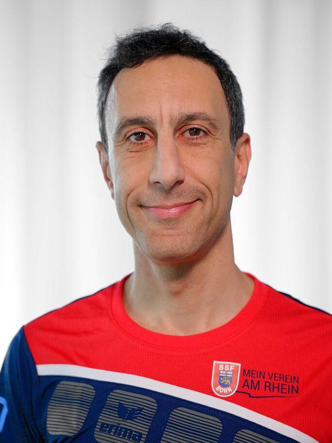 Porträt eines Mannes in einem rot-blauen Sporttrikot mit dem Schriftzug „Mein Verein am Rhein“ vor neutralem Hintergrund.