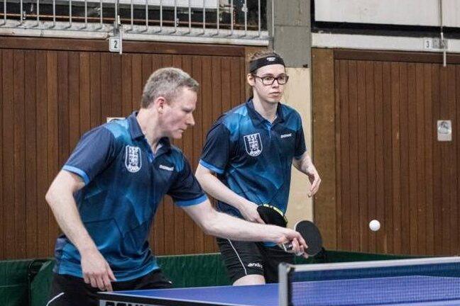 Zwei Tischtennisspieler in blauen Trikots bei einem Match an einem Tischtennistisch, Ball in der Luft.