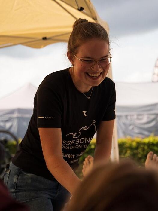 Eine lächelnde Frau in einem T-Shirt steht unter einem Zelt und gibt einer Person eine Massage.