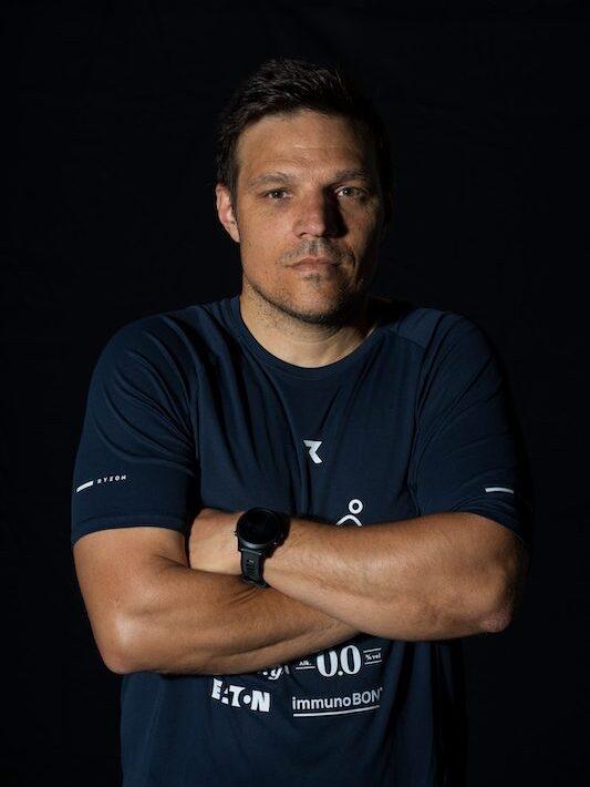 Ein sportlicher Mann mit kurzen Haaren steht mit verschränkten Armen vor einem schwarzen Hintergrund.