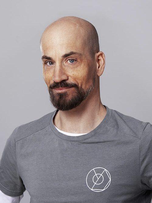 Mann mit Vollem Bart und Glatze lächelt, trägt ein graues T-Shirt mit weißem Logo, vor grauem Hintergrund.