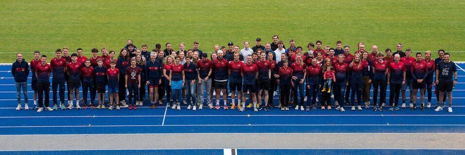 Gruppenfoto von Sportlern in roten Shirts auf einer blauen Laufbahn im Freien, umgeben von grünem Rasen.