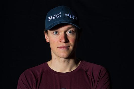 Portrait eines Mannes mit verschränkten Armen, trägt ein rotes T-Shirt des SSF Bonn Triathlon Teams und eine blaue Cap.