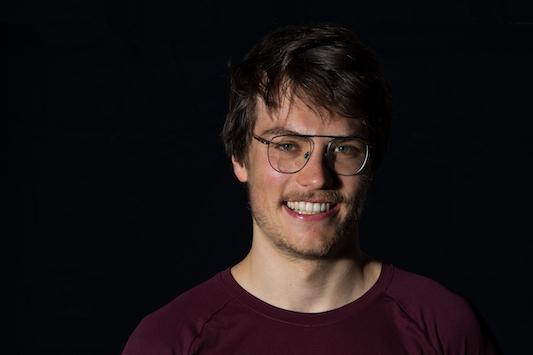 Lächelnder Mann mit Brille trägt ein rotes T-Shirt des SSF Bonn Triathlon Teams vor schwarzem Hintergrund.