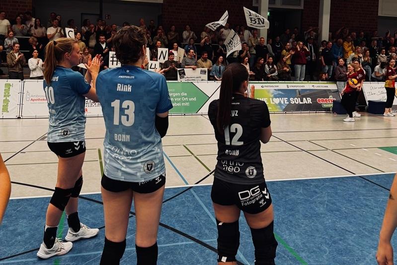 Drei Volleyballerinnen in Trikots stehen an der Spielfeldlinie, während eine jubelnde Menge im Hintergrund das Spiel unterstützt.