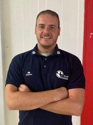 Lächelnder Mann in Sportbekleidung mit verschränkten Armen vor einer farbigen Wand.