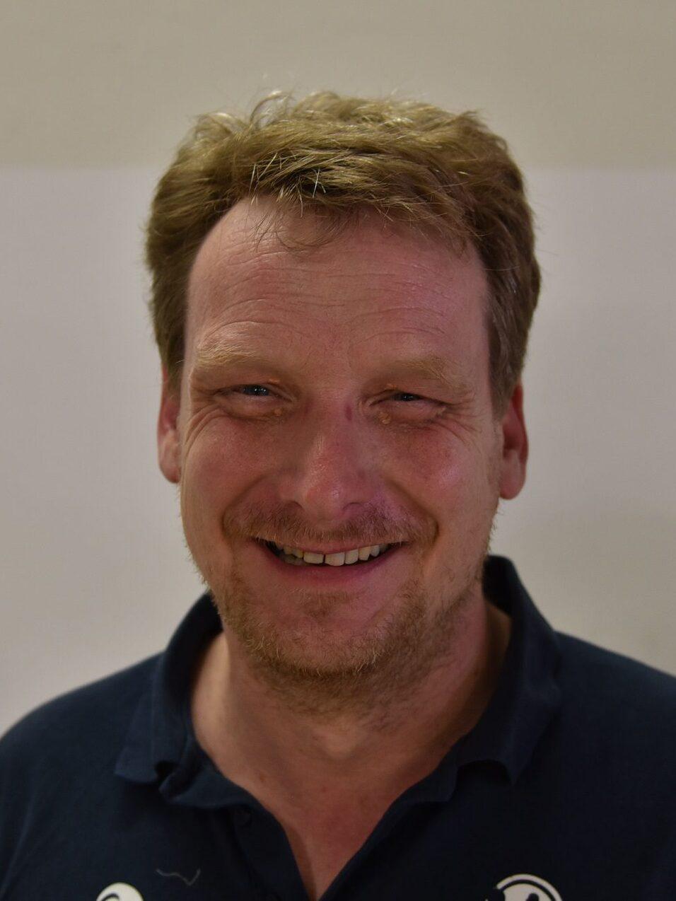 Porträt eines lächelnden Mannes mit kurzen, blonden Haaren, in einem dunklen Polohemd vor einer hellen Wand.