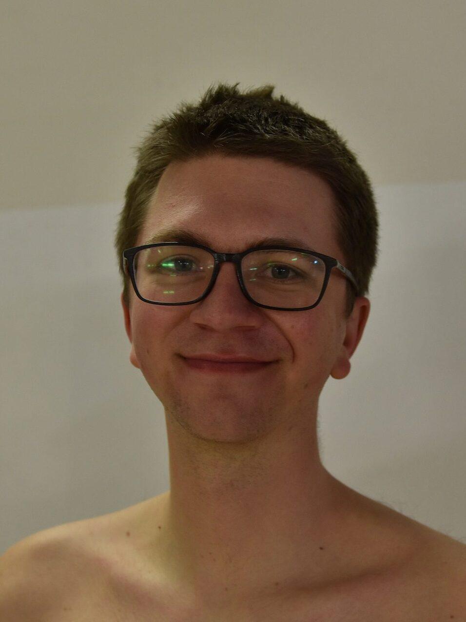Junger Mann mit Brille, lächelnd vor einfarbiger Wand, ohne Oberbekleidung.