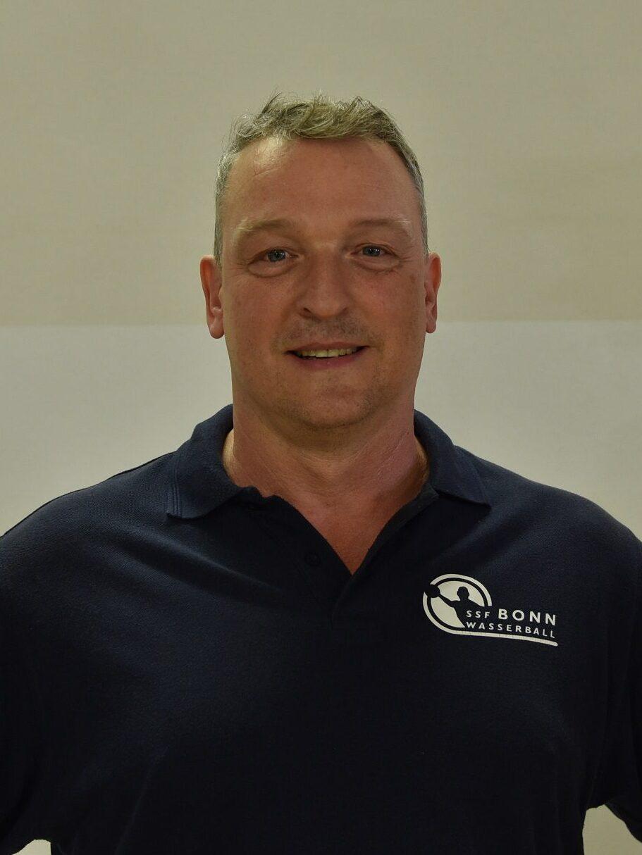 Mann in blauem Polo-Shirt mit Logo steht vor einer hellen Wand und lächelt in die Kamera.