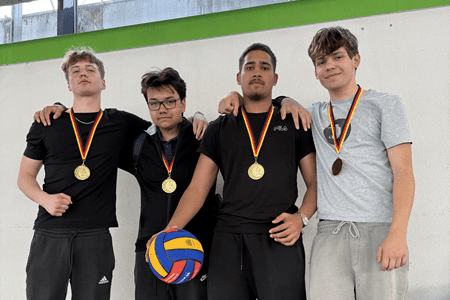 Vier Jugendliche in sportlichen Outfits stehen gemeinsam, jeder trägt eine Medaille und hält einen Volleyball.