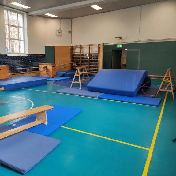 Sporthalle mit Turngeräten, blauen Matten und Barren auf einem grünen Boden, hell erleuchtet von großen Fenstern.