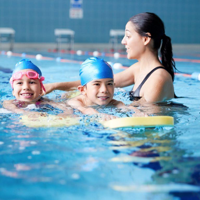 Zwei Kinder schwimmen im Wasser, während eine Schwimmtrainerin sie mit einem Schwimmbrett unterstützt.