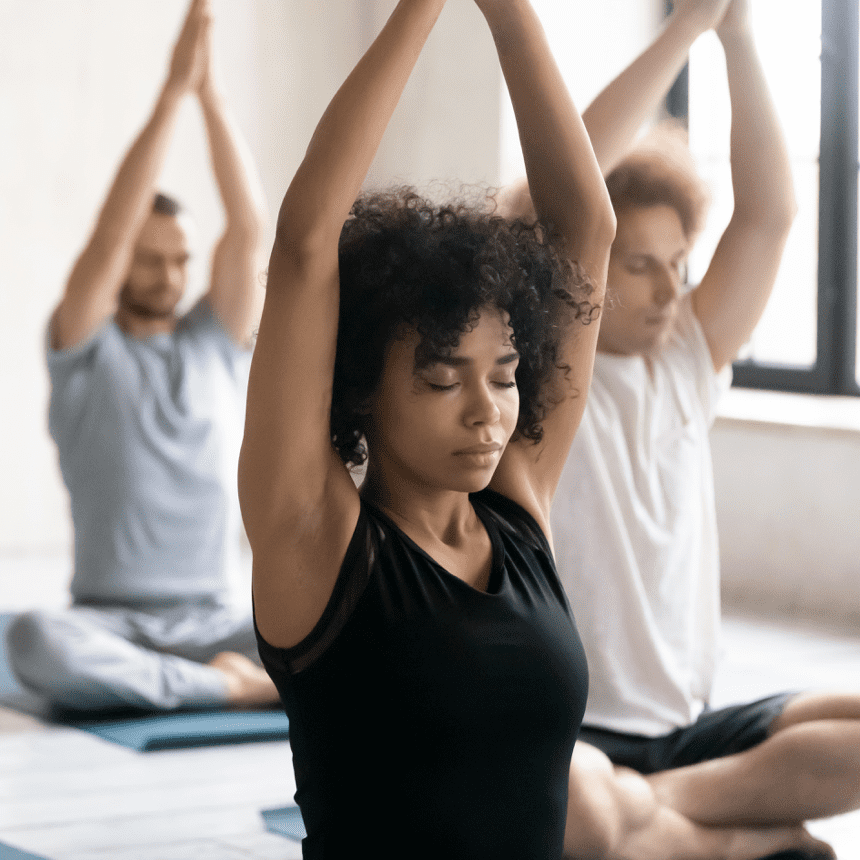 Drei Menschen praktizieren Yoga in einem hellen Raum; eine Frau mit lockigem Haar hält die Hände über dem Kopf.