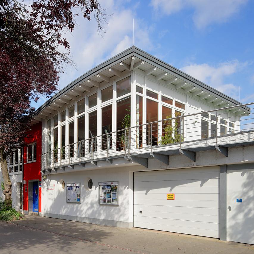 Moderner Bau mit großen Fenstern, roten und weißen Fassaden, umgeben von Bäumen und einem Gehweg.