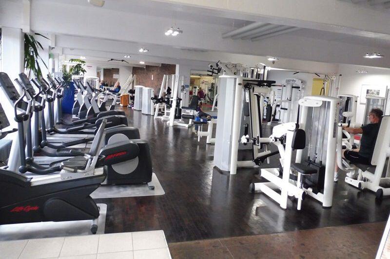 Fitnessstudio mit mehreren Trainingsgeräten und Ausdauermaschinen, während Nutzer aktiv trainieren. Helle, geräumige Umgebung.