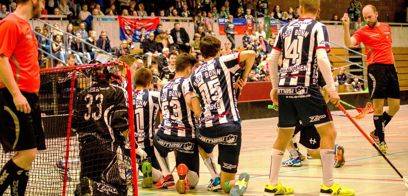 Szenario eines Floorball-Spiels mit Spielern in Schwarz und Weiß vor Tor und jubelnden Zuschauern im Hintergrund.