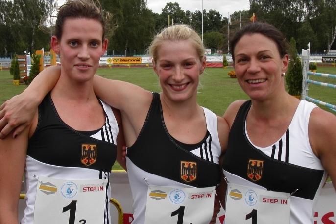 Drei Athletinnen in Sportkleidung stehen eng beieinander und lächeln vor einer Wettkampfstätte für Modernen Fünfkampf.